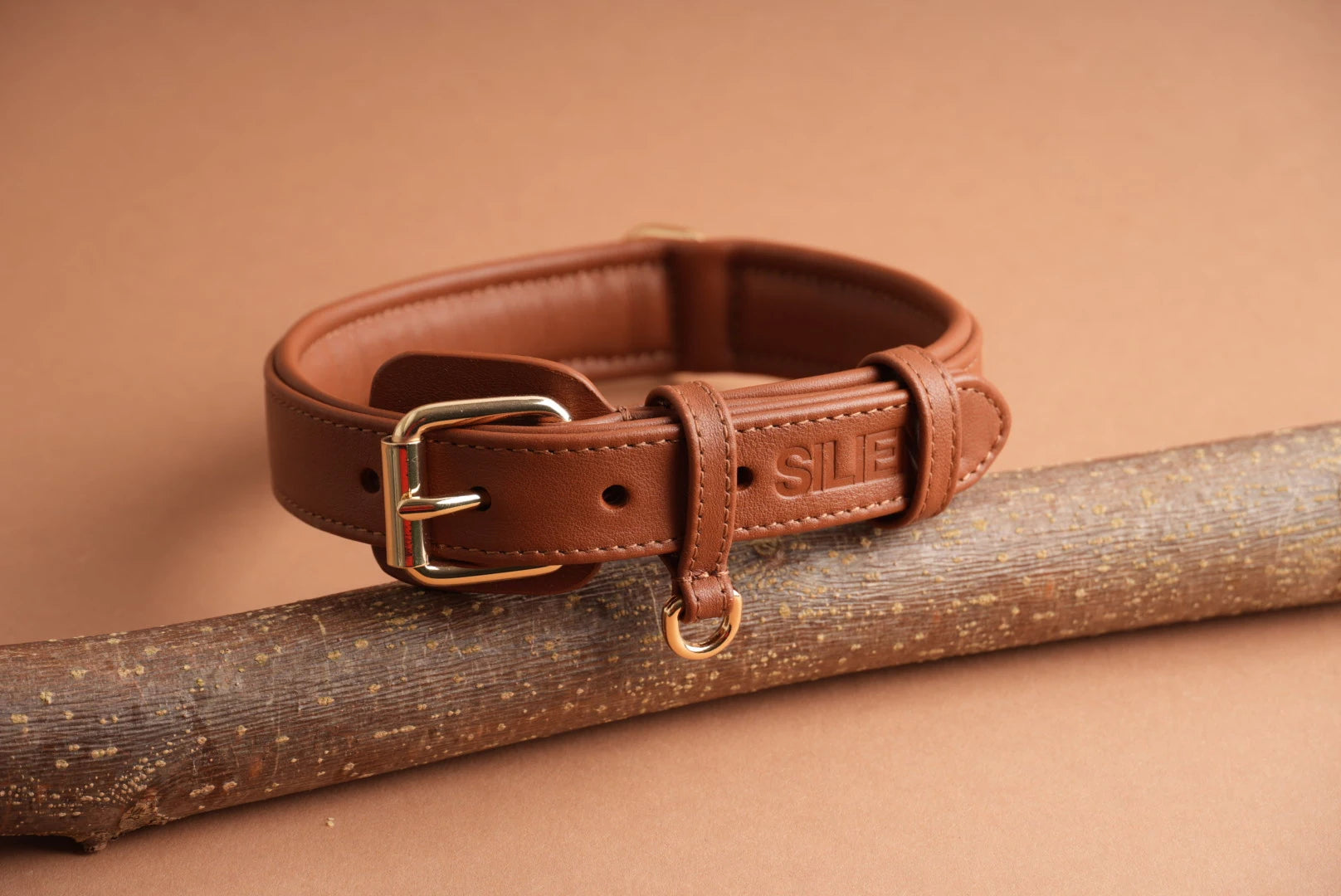 Hundehalsband aus braunem Echtleder von Siliena mit goldener Schnalle, handgefertigt und gepolstert, auf einem Ast drapiert vor beigem Hintergrund.